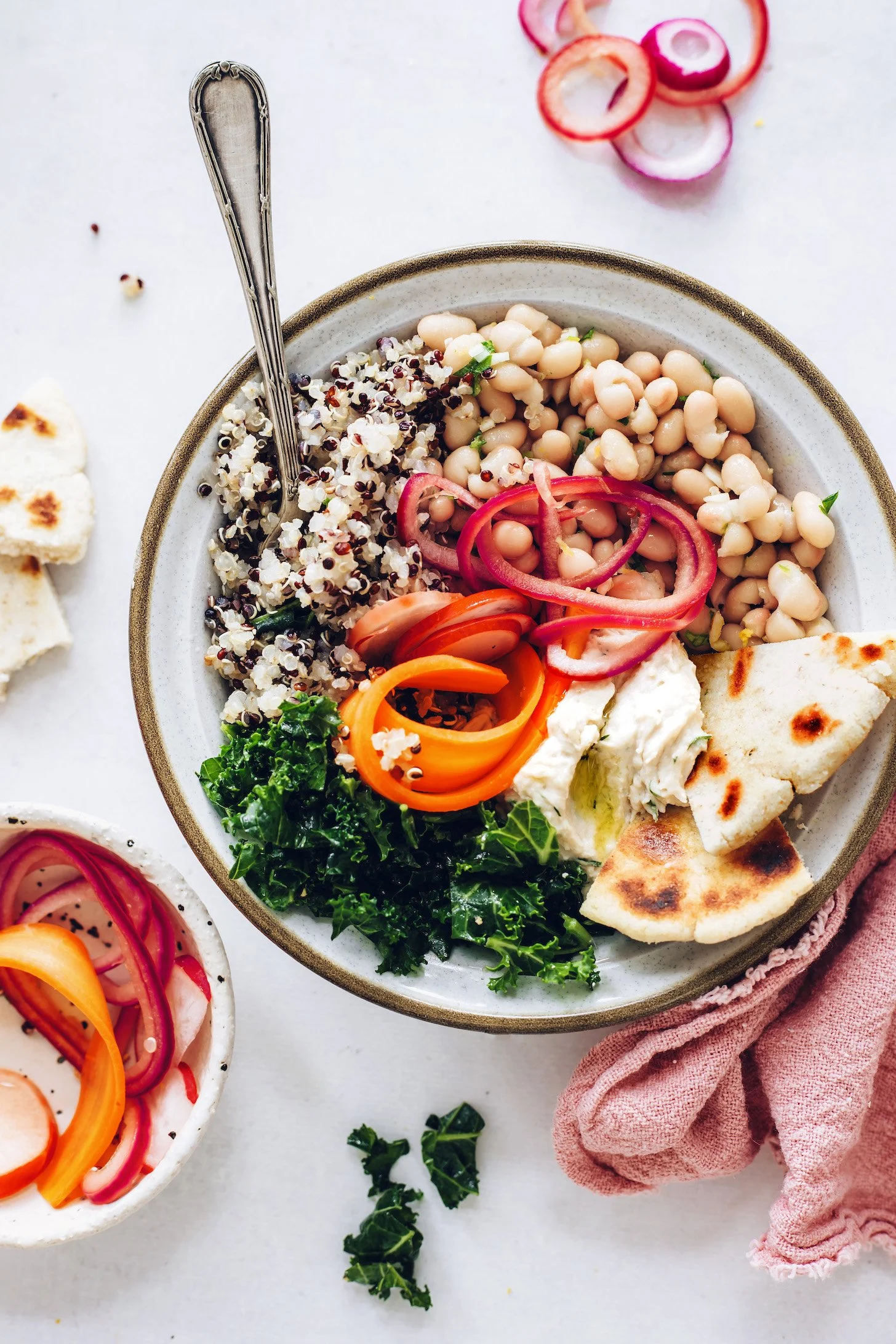 A bowl of mixed salad with quinoa, chickpeas, sliced tomatoes, peppers, and greens, accompanied by pita bread and onion slices on a white surface.