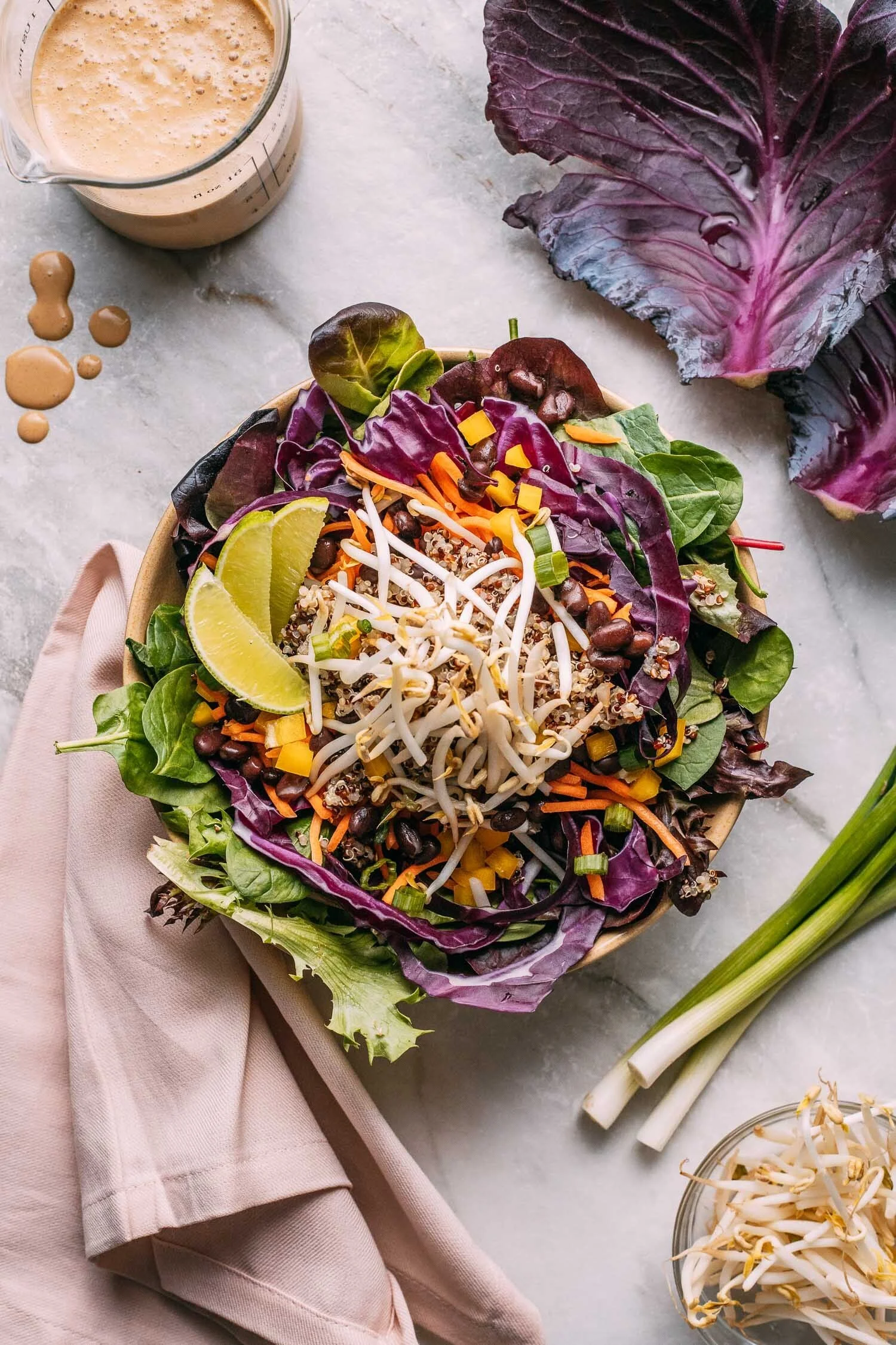 A bowl of fresh salad with mixed greens, purple and green lettuce, shredded carrots, black beans, diced yellow peppers, bean sprouts, and lemon wedges, on a marble surface with salad ingredients and dressing nearby.