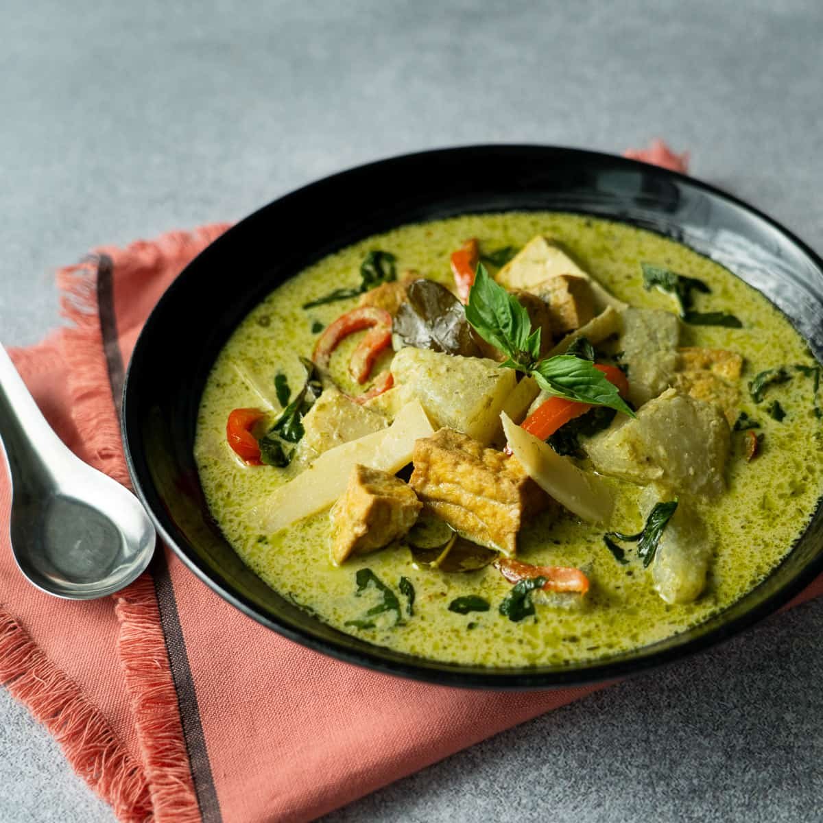 A bowl of vegan green curry with vegetables and chunks of tofu garnished with basil leaves, served on a pink napkin with a spoon.