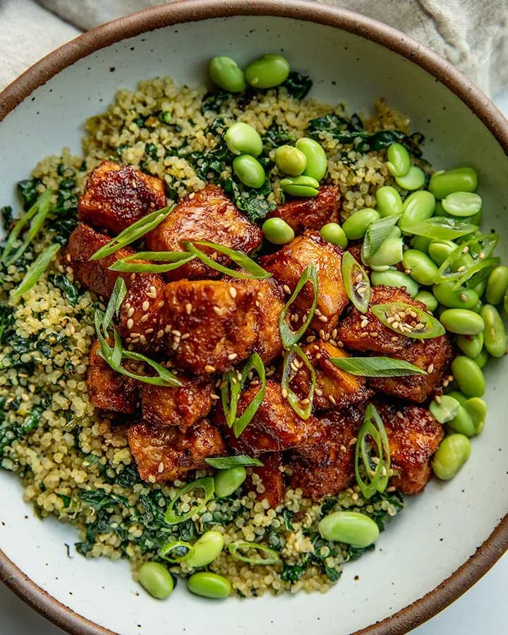 A bowl of cooked quinoa topped with tofu pieces, sliced green onions, edamame, and chopped kale.