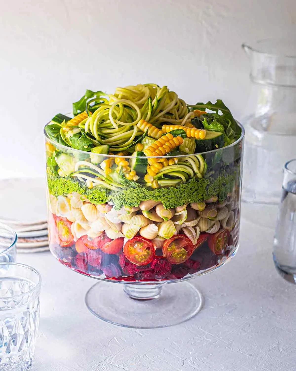 Layered salad in a large glass bowl with vegetables, pasta, and cherry tomatoes, on a white table with glasses of water nearby.