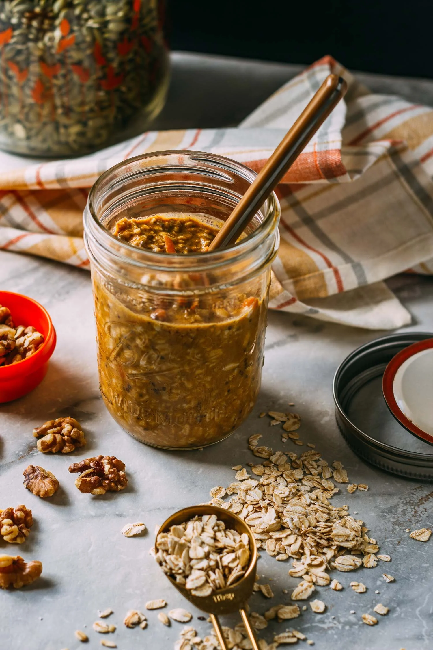 A jar of oatmeal with a metal straw, surrounded by scattered oats, walnuts, and a small red bowl of walnuts, with a black and white checkered dish towel on a marble surface.