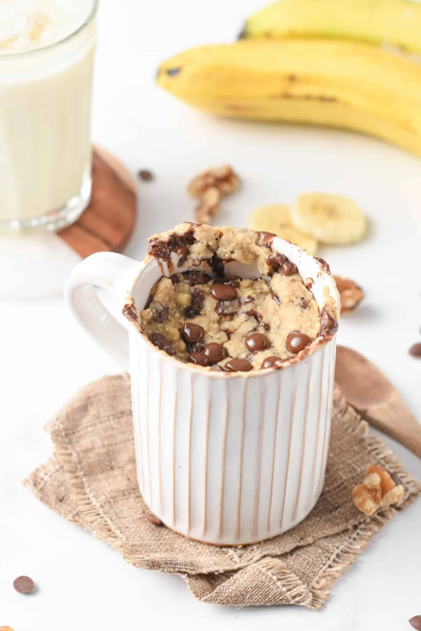 Chocolate chip cookie dough in a mug with spilled chocolate chips, bananas, walnuts, and a glass of banana smoothie in the background.