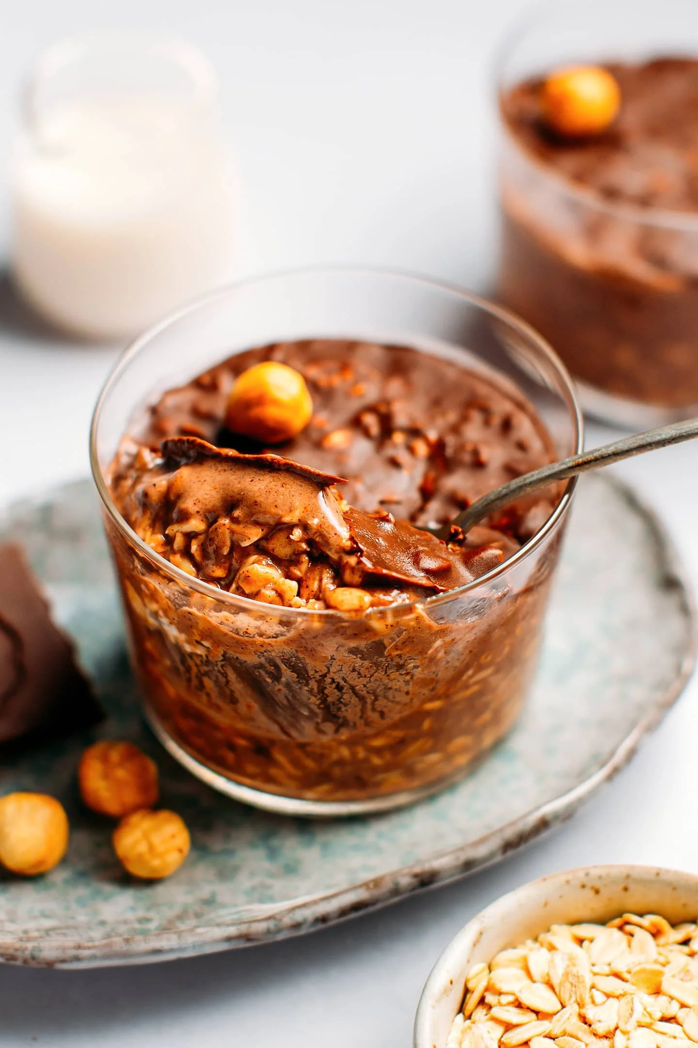 Glass bowl of chocolate mousse topped with hazelnuts on a ceramic plate with hazelnuts, chocolate, and a glass of milk in the background.