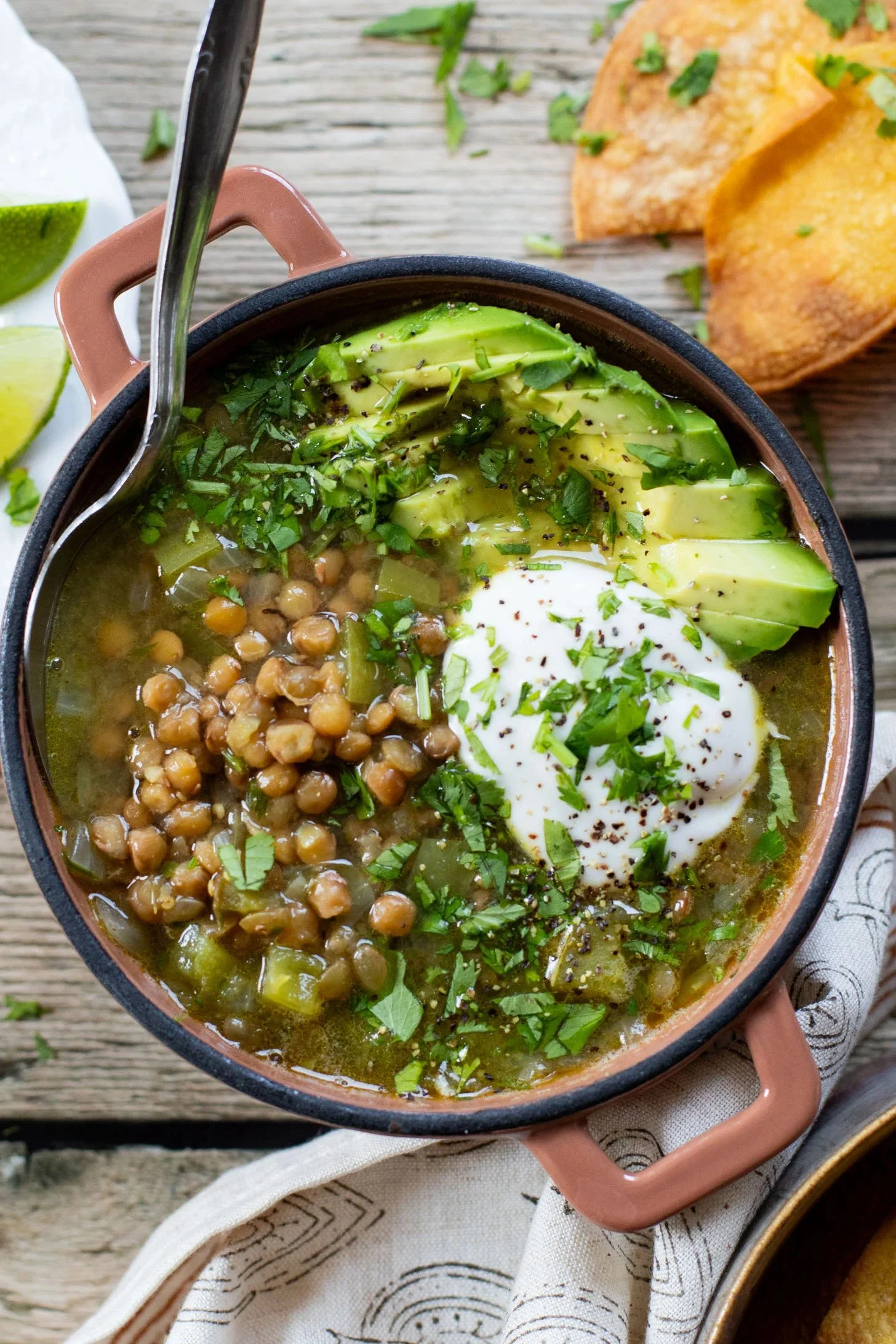 A bowl of veggie and chickpea soup topped with sliced avocado, fresh cilantro, a dollop of sour cream, and black pepper, with crispy tortilla chips on the side.