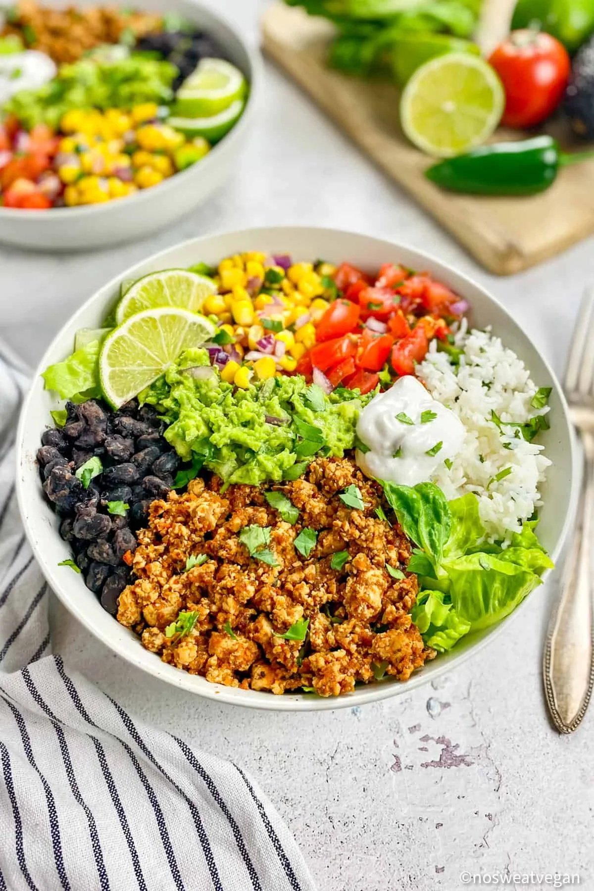 A colorful vegan burrito bowl with rice, black beans, seasoned tofu, guacamole, chopped tomatoes, corn, chopped red onions, sour cream, and lime slices, on a white surface with a striped napkin.