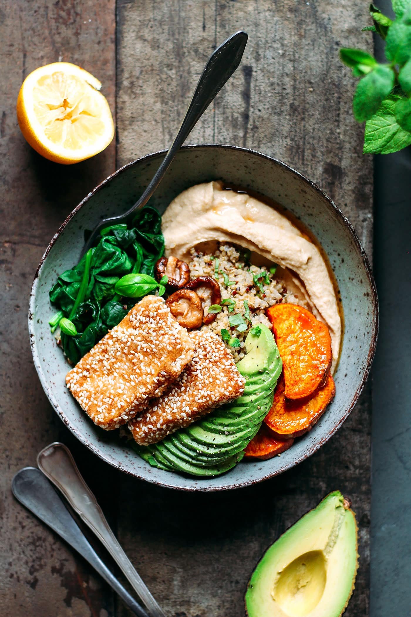 A bowl of healthy food containing sautéed greens, fried tofu, sliced avocado, roasted sweet potatoes, a scoop of hummus, and quinoa, with half a lemon, a sliced avocado, and green herbs nearby on a rustic wooden surface.