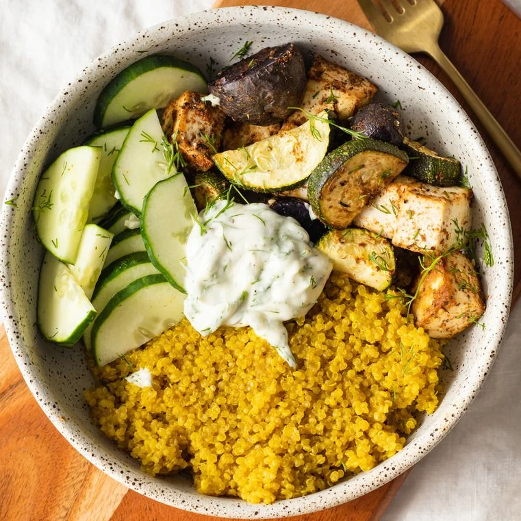 A bowl of Mediterranean-inspired food with yellow couscous, grilled chicken pieces, sliced cucumbers, roasted zucchini and eggplant, garnished with fresh herbs and a dollop of tzatziki sauce.