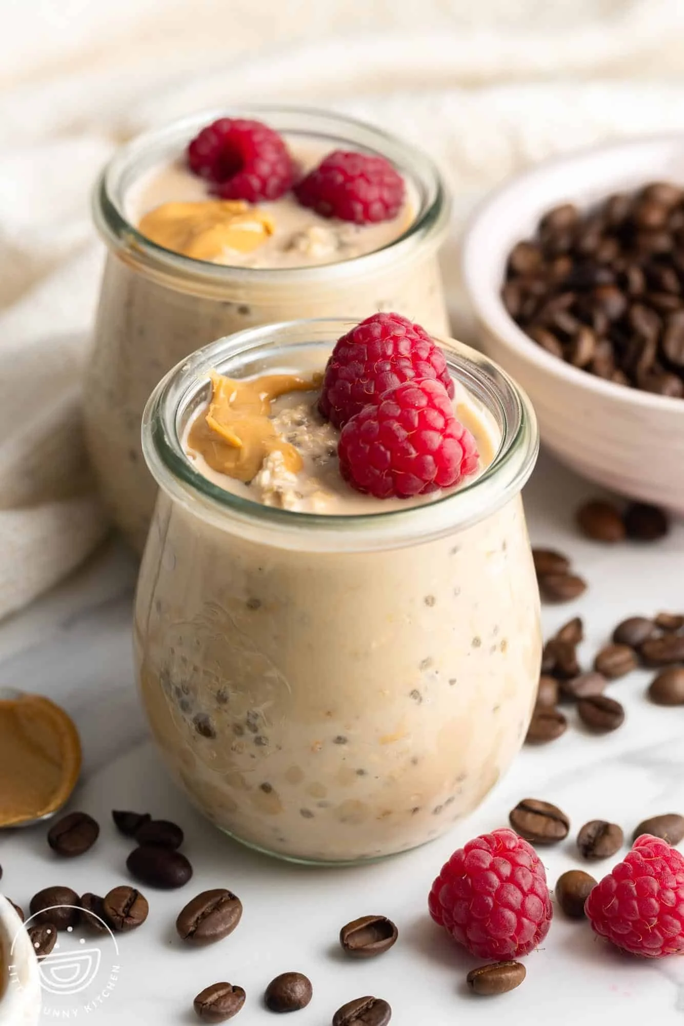 Two glasses of creamy overnight oats topped with fresh raspberries and drizzled with peanut butter, surrounded by coffee beans and a bowl of more coffee beans, on a white surface.