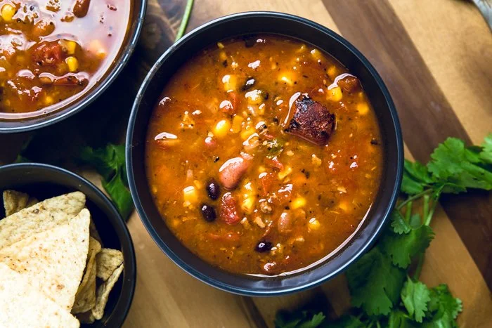 A bowl of chili with various beans, corn, and a piece of meat, placed on a wooden surface with cilantro leaves nearby.