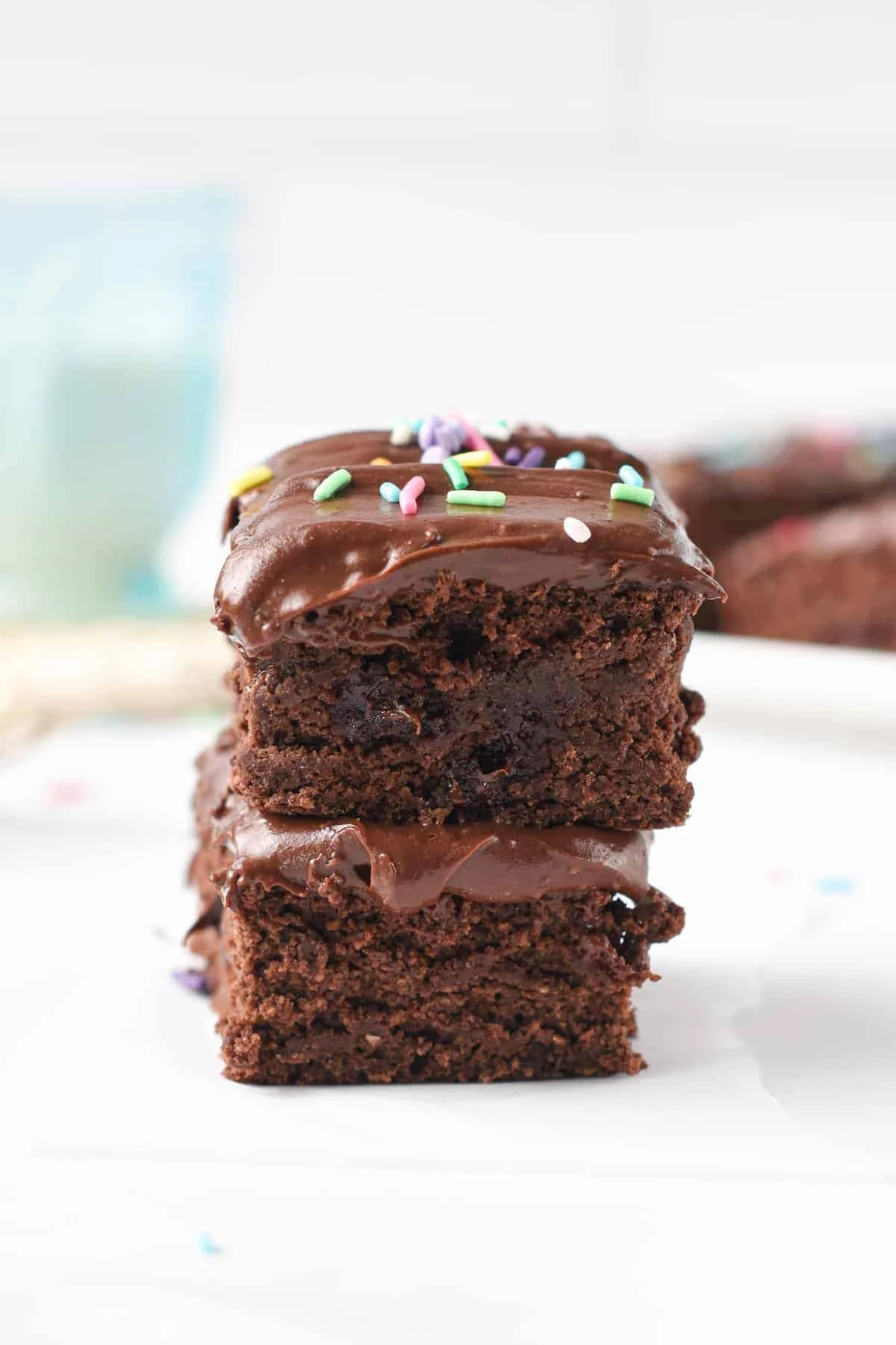 Two stacked chocolate brownies with glossy chocolate frosting and colorful sprinkles on top, on a white surface.
