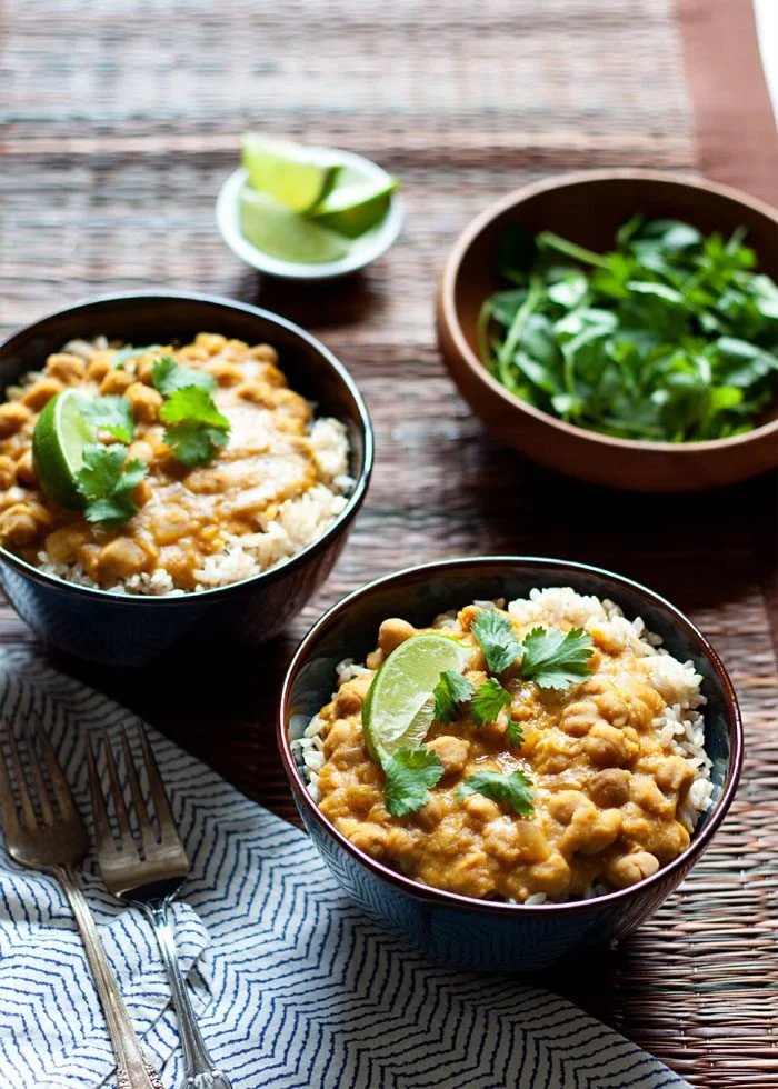 Two bowls of chickpea curry with rice, garnished with lime wedges and cilantro, on a woven wooden table with a small bowl of salad and lime wedges in the background.