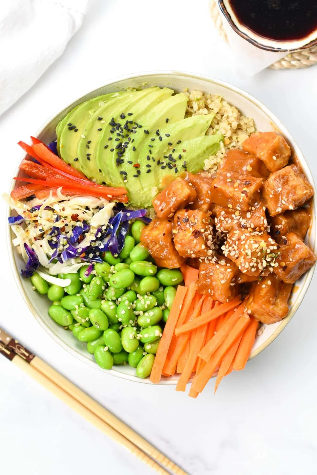 A colorful poke bowl with avocado slices, tofu in sauce, shredded cabbage, shredded carrots, edamame, quinoa, and red peppers, topped with sesame seeds, with a glass of dark soda nearby.