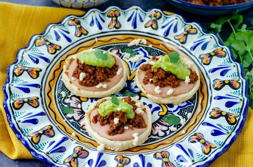 Three mini vegan sopas with ground walnut meat, guacamole, and white cotija vegan cheese on a colorful ceramic plate with intricate blue, yellow, and white patterns, placed on a yellow cloth with green cilantro leaves nearby.