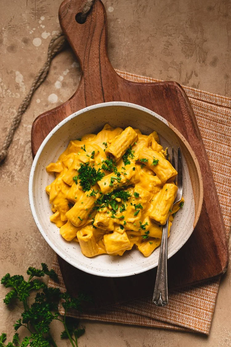 A bowl of creamy pasta with yellow cheese sauce, garnished with chopped parsley, on a wooden serving board with a fork.