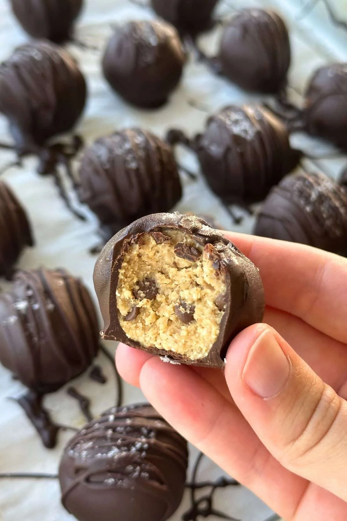 Close-up of a hand holding a bitten vegan chocolate caramel truffle with a soft caramel center and chocolate coating, with a background of several more unbitten truffles.