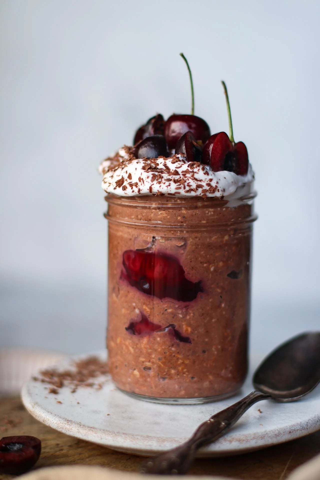 Chocolate dessert in a glass jar topped with whipped cream, cherries, and chocolate shavings, with a spoon on a white plate.
