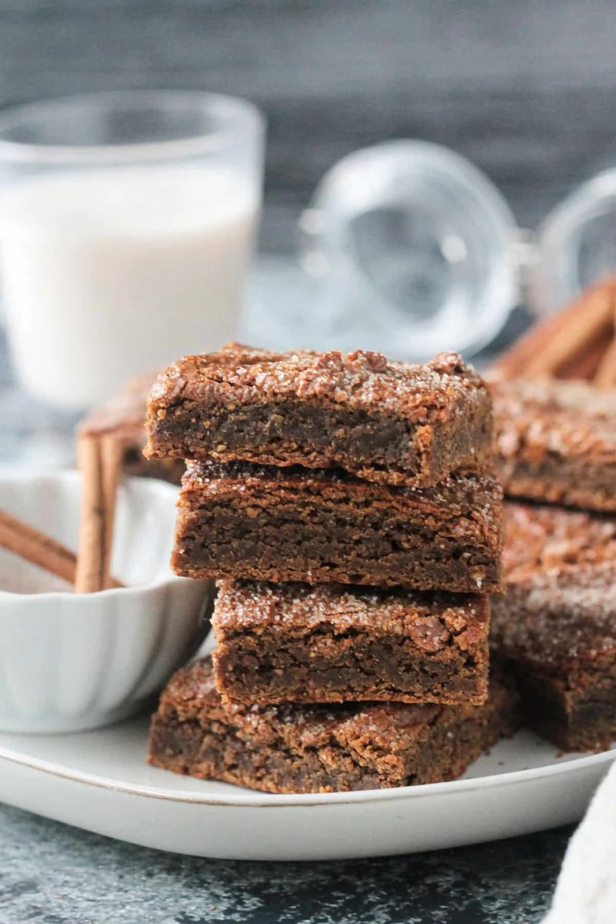 Stack of four vegan chocolate brownies on a white plate, with a small bowl of sugar next to them and a glass of milk in the background.