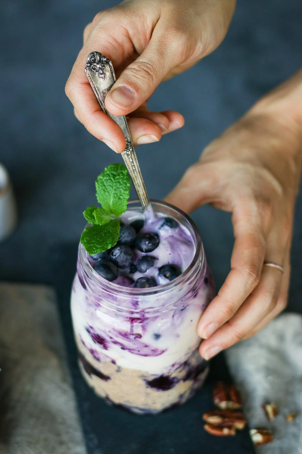 A person is holding a spoon over a mason jar filled with blueberries, whipped cream, and purple berry sauce, garnished with a sprig of mint.