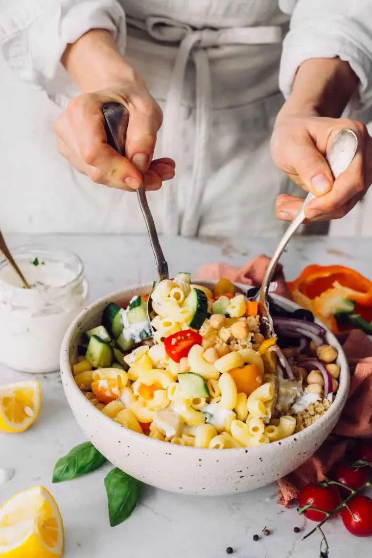 Person adding pasta, chickpeas, cucumbers, and cherry tomatoes to a large salad bowl with a spoon.