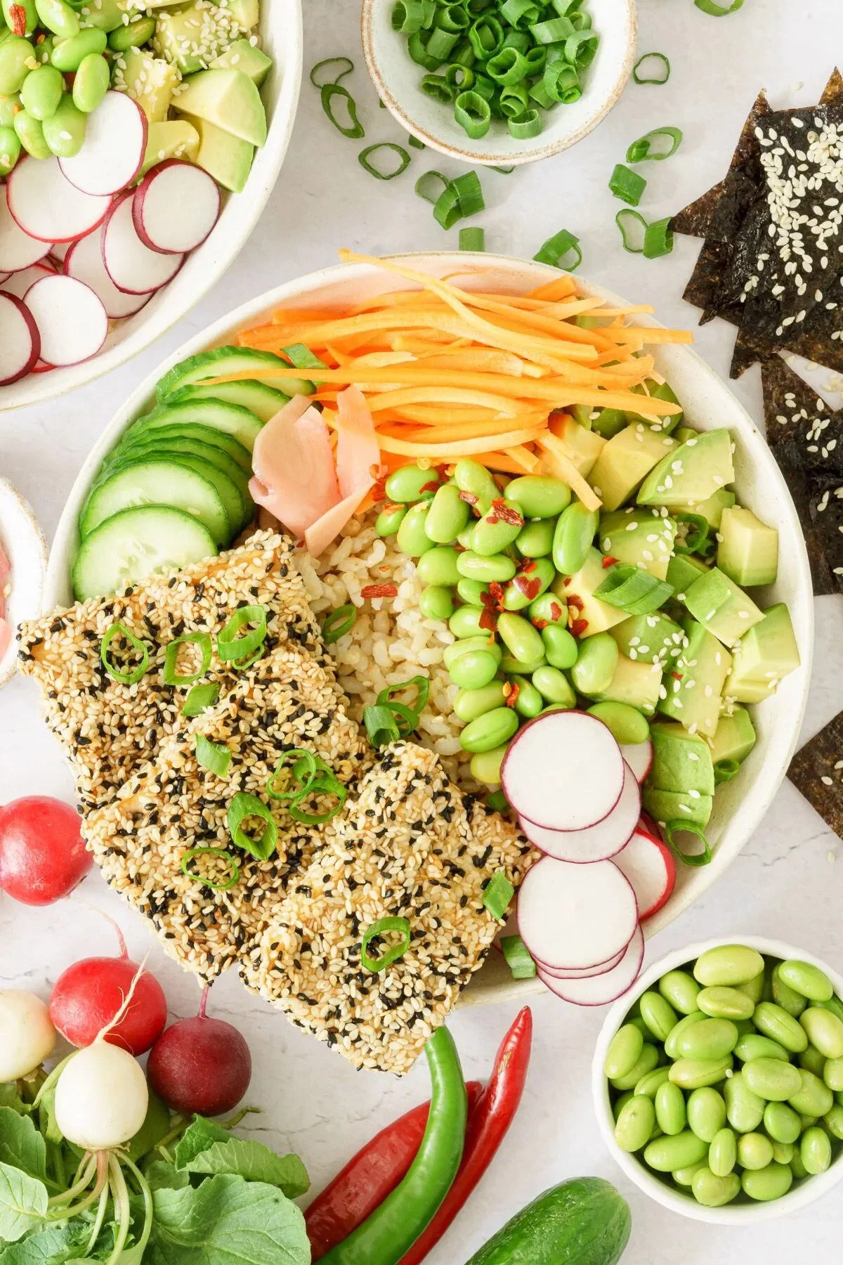 A colorful poke bowl containing sliced cucumber, shredded carrots, diced avocado, edamame, pickled ginger, rice, and sesame-crusted tofu garnished with green onions, surrounded by small bowls of chopped green onions and edamame, and fresh radishes and chili peppers.