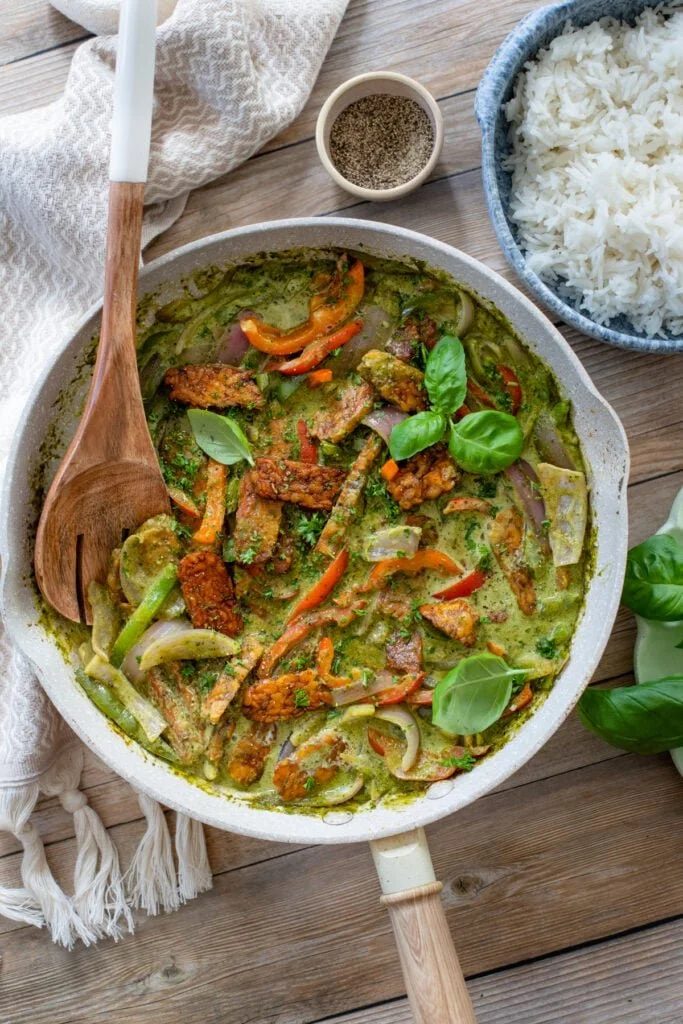 A skillet with a green curry dish containing vegetables and pieces of fried tofu, garnished with fresh basil leaves, served with a bowl of white rice and a small dish of black pepper on a wooden table.