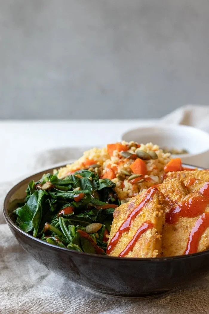 A black bowl containing fried fish fillets with ketchup, sautéed greens with pumpkin seeds, and mashed sweet potatoes with carrots and herbs.