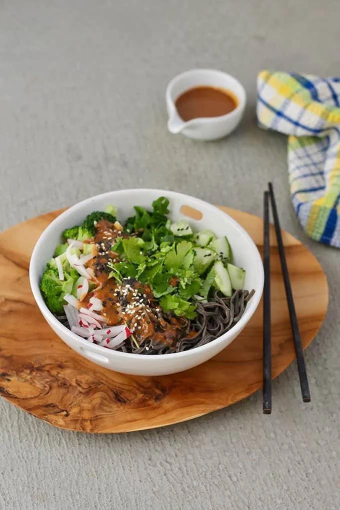 Bowl of Asian noodle salad with broccoli, cucumbers, radishes, green onions, cilantro, and black noodles, topped with sesame seeds and sauce, on a round wooden serving board. Small bowl of sauce and black chopsticks beside the bowl.