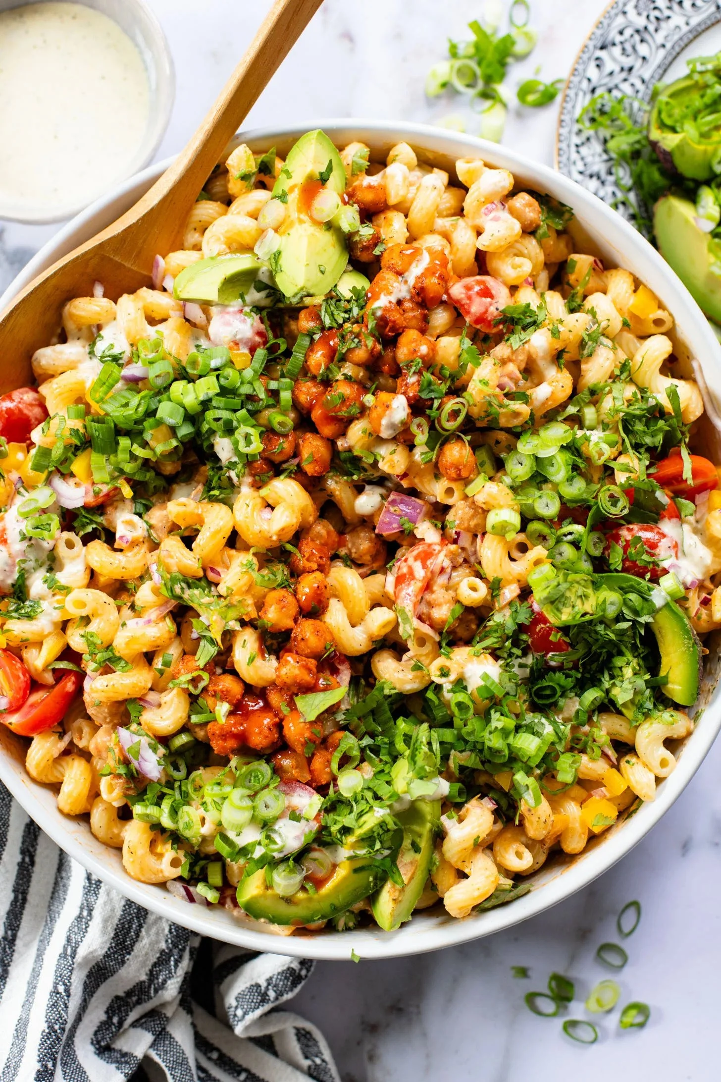 Bowl of vegan macaroni salad topped with chopped green onions, cilantro, avocado slices, cherry tomatoes, and a sprinkle of dressing, with a side of salad and a bottle of dressing.