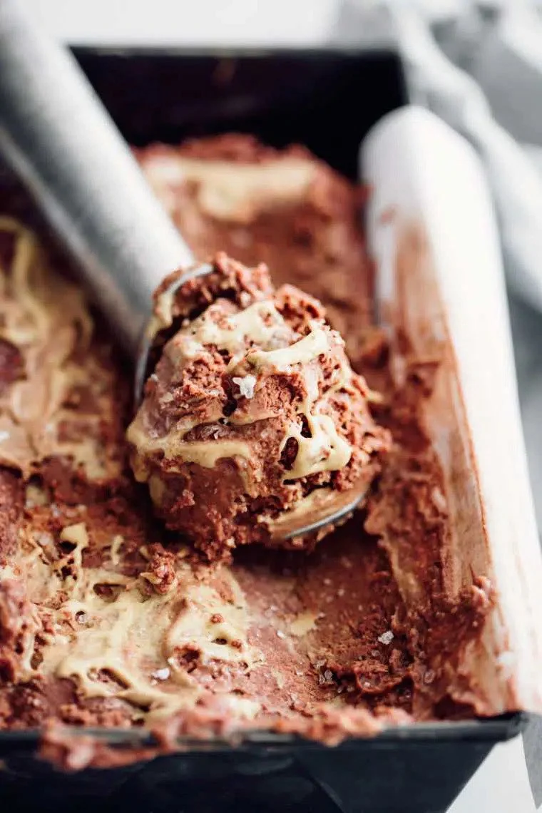 Close-up of partially eaten vegan chocolate ice cream with chunks in a black container, with an ice cream scoop resting on top.