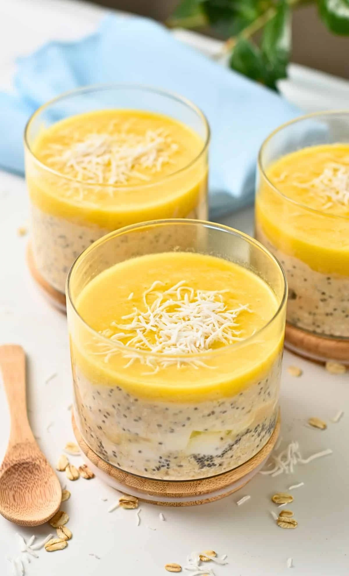 Three glasses of layered overnight oats topped with lemon curd and shredded cheese, with a blue cloth napkin and scattered oats in the background.