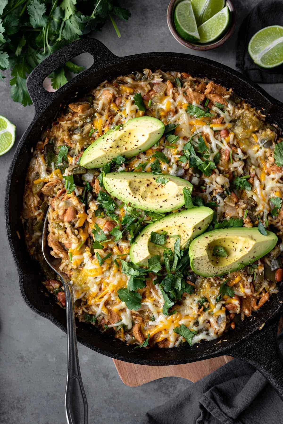 A cast iron skillet filled with cheesy taco beef rice casserole topped with sliced avocado, cilantro, and sprinkled with black pepper, with lime wedges and lime slices on the side.