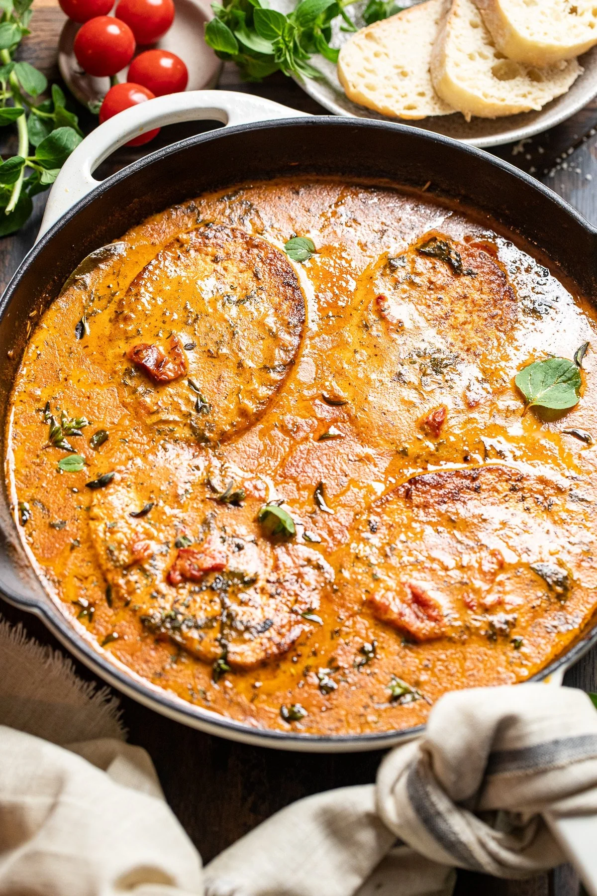 A skillet of tofu with tomato-based sauce, garnished with herbs, alongside a plate of sliced bread and cherry tomatoes.