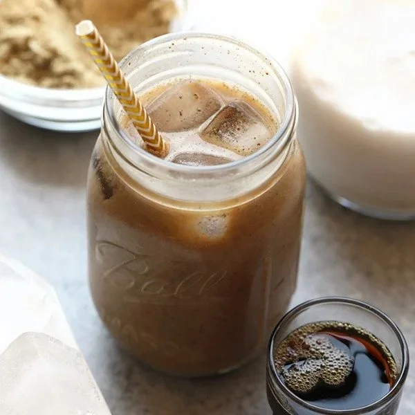 A mason jar filled with iced coffee and ice cubes, with a yellow and white striped straw, on a light-colored surface alongside smaller glasses of dark soda and additional beverages.
