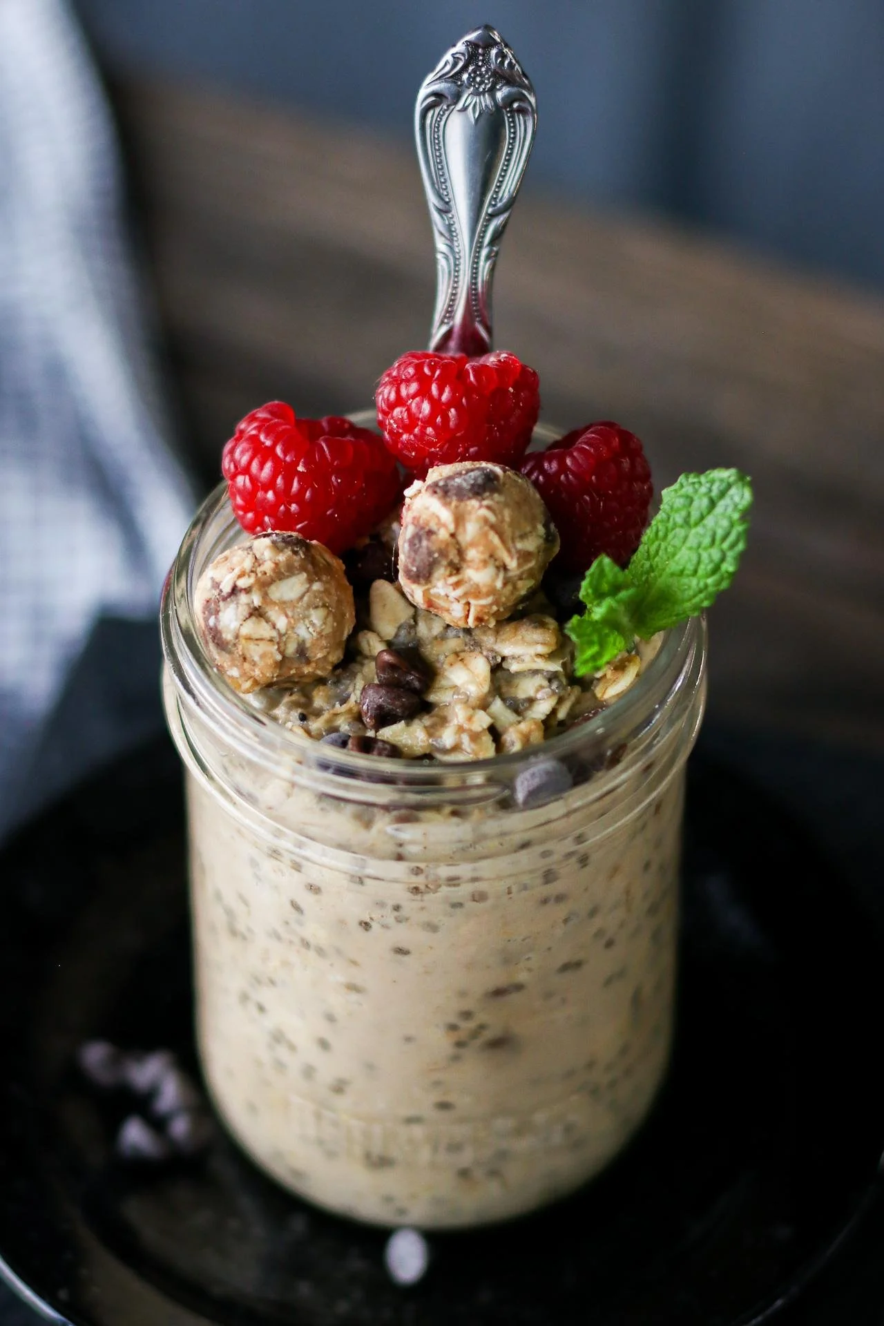 A glass jar filled with overnight oats topped with fresh raspberries, chocolate chips, granola balls, a mint leaf, and a spoon.