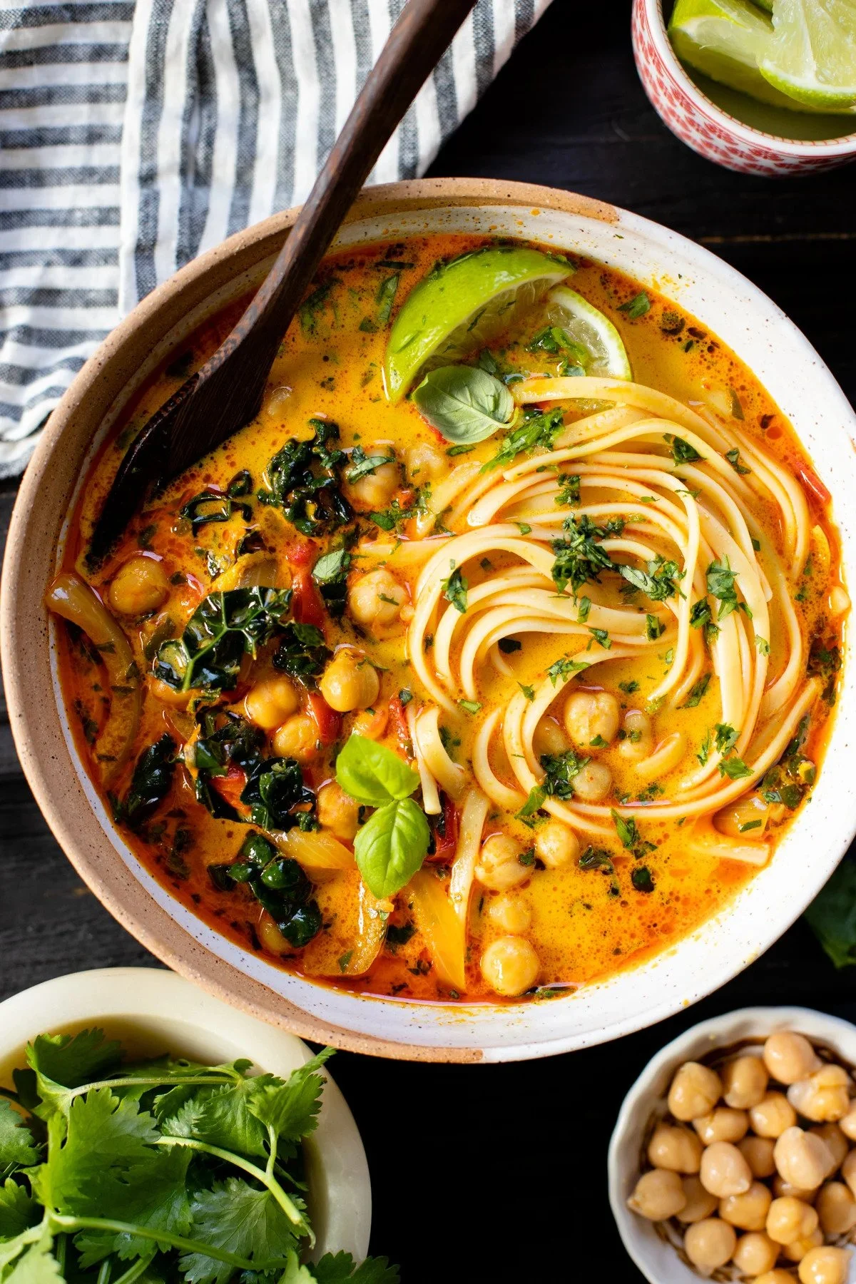 A bowl of vegan chickpea and vegetable curry with noodles, garnished with cilantro, basil, and lime wedges, with side bowls of chickpeas and cilantro on a dark table.