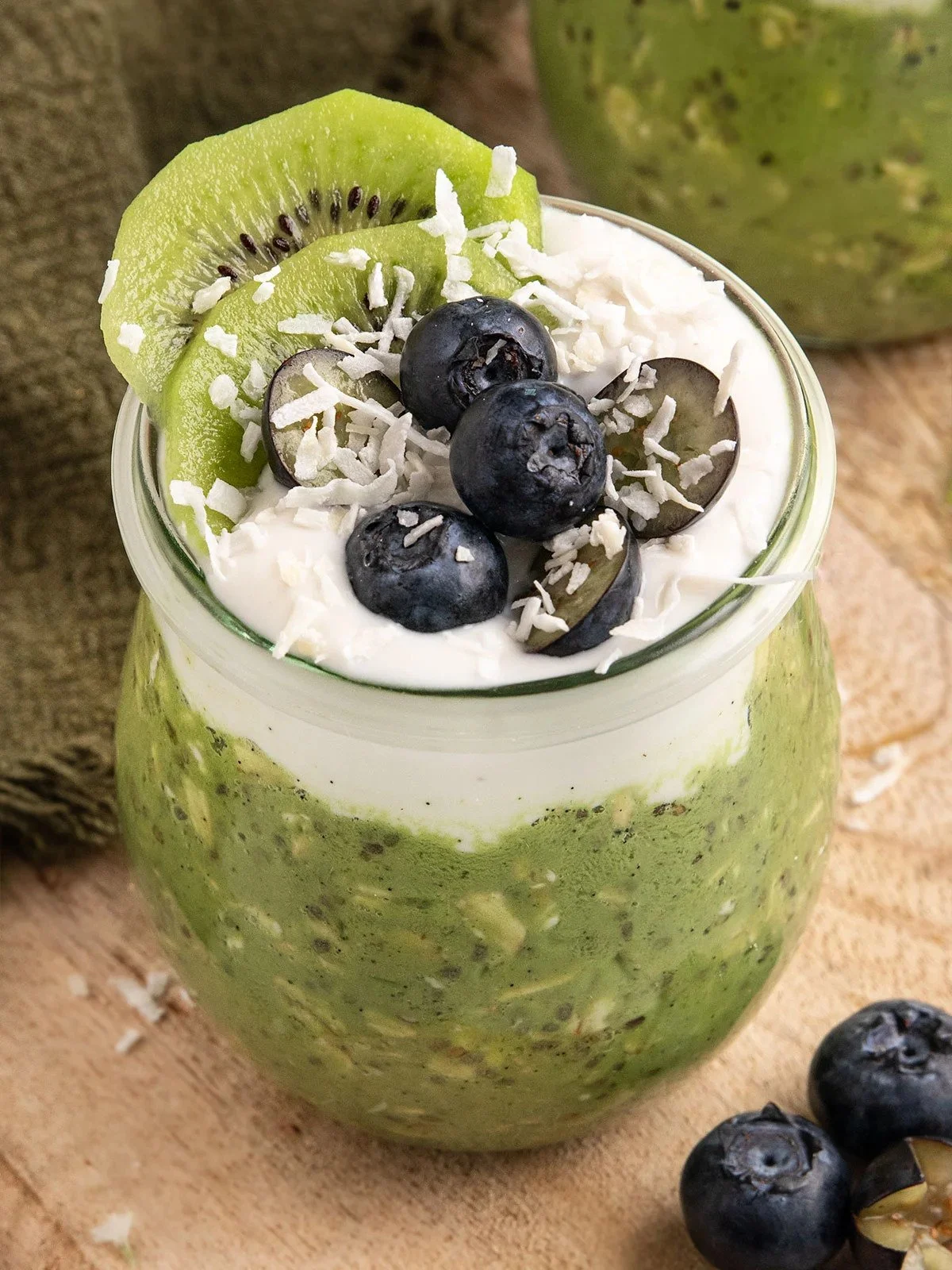 A glass jar filled with a green smoothie topped with slices of kiwi, blueberries, and shredded coconut.