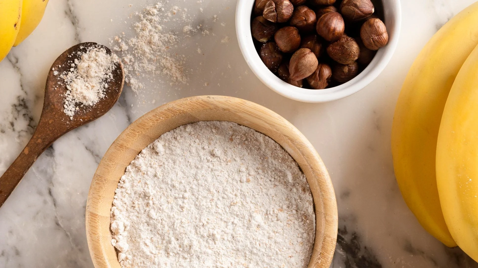 A marble countertop with ingredients including a wooden bowl of vegan protein powder, a white bowl of hazelnuts, a banana, and a wooden spoon with protein powder on it.