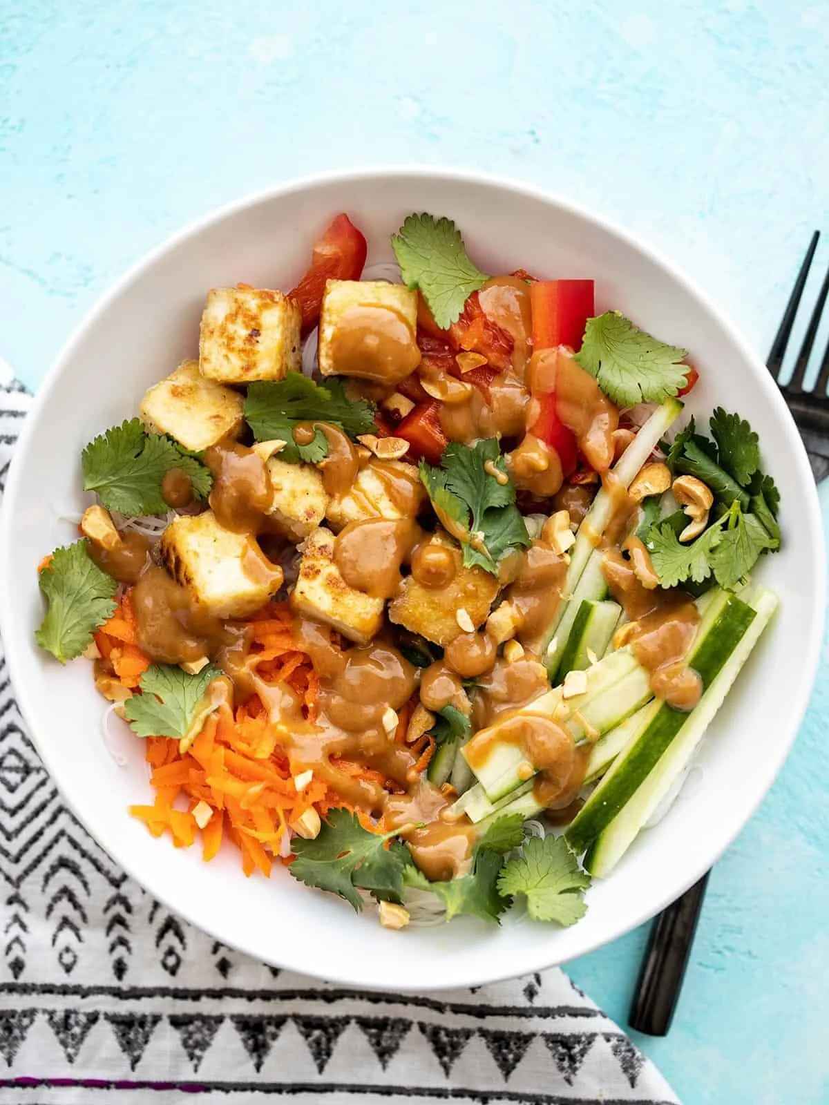 Buddha bowl with grilled tofu, shredded carrots, cherry tomatoes, cucumber sticks, cilantro, peanuts, and peanut sauce.