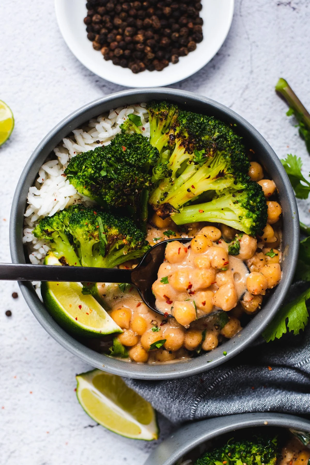 A bowl of rice, grilled broccoli, and chickpea stew garnished with lime wedges on a gray surface.