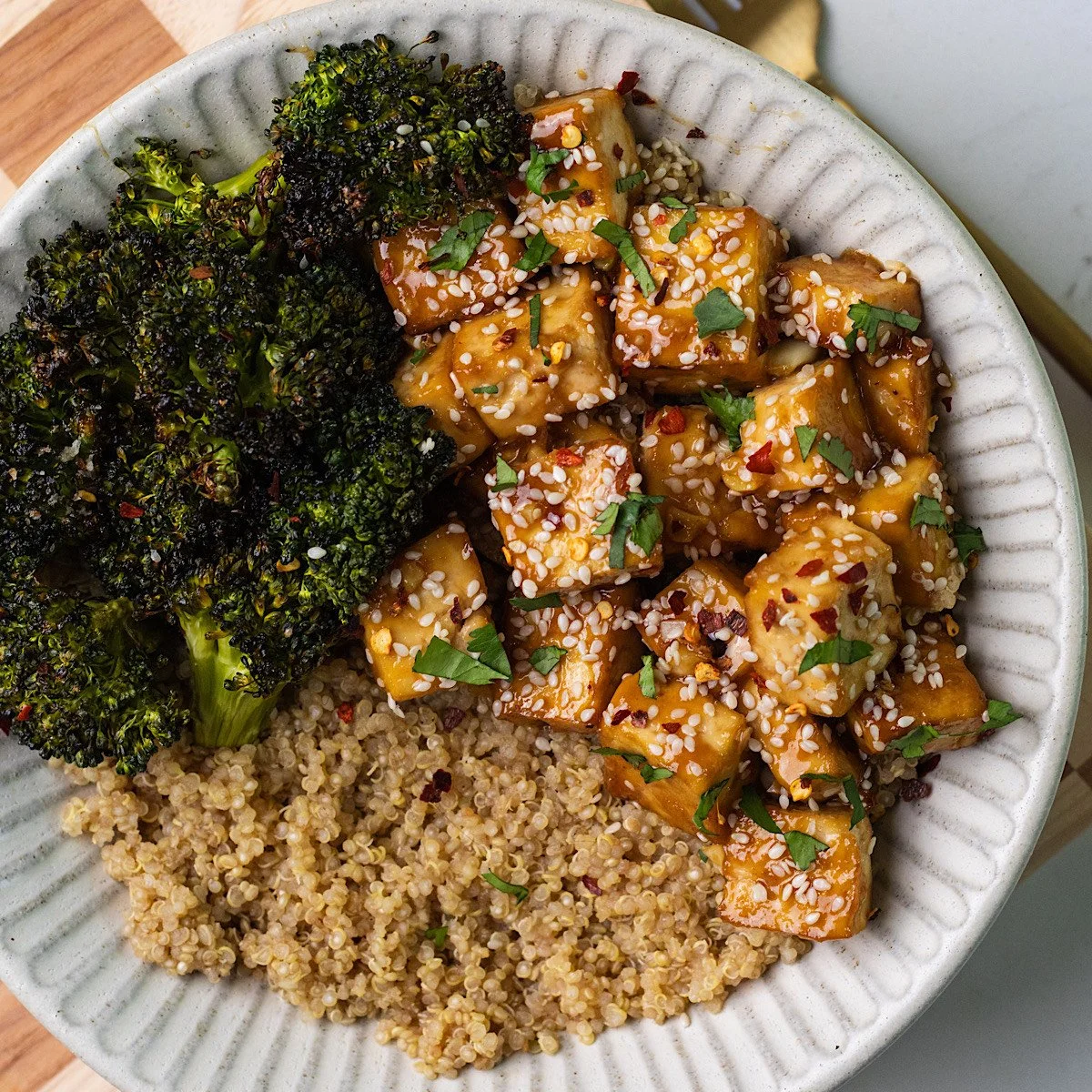A bowl of roasted broccoli, cooked quinoa, and tofu cubes glazed in sauce, garnished with sesame seeds and chopped herbs