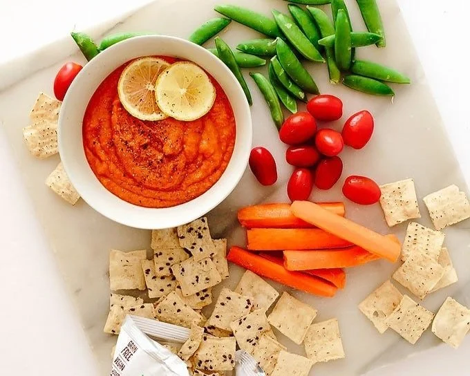 Fresh vegetables and a bowl of red hummus on a white platter. The vegetables include cherry tomatoes, julienned carrots, snap peas, and cubed pita chips.