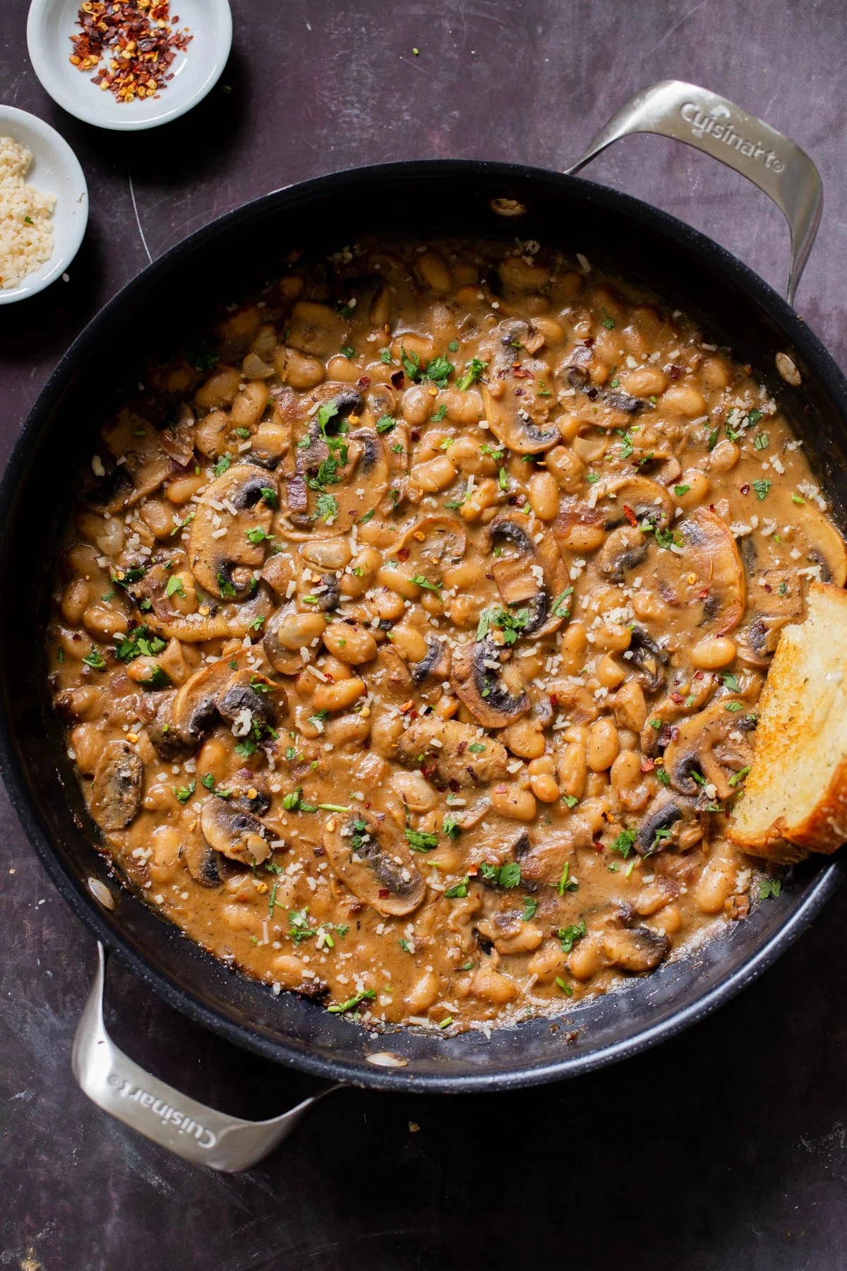 Creamy mushroom and bean stew garnished with parsley in a black skillet with a piece of bread on the side.
