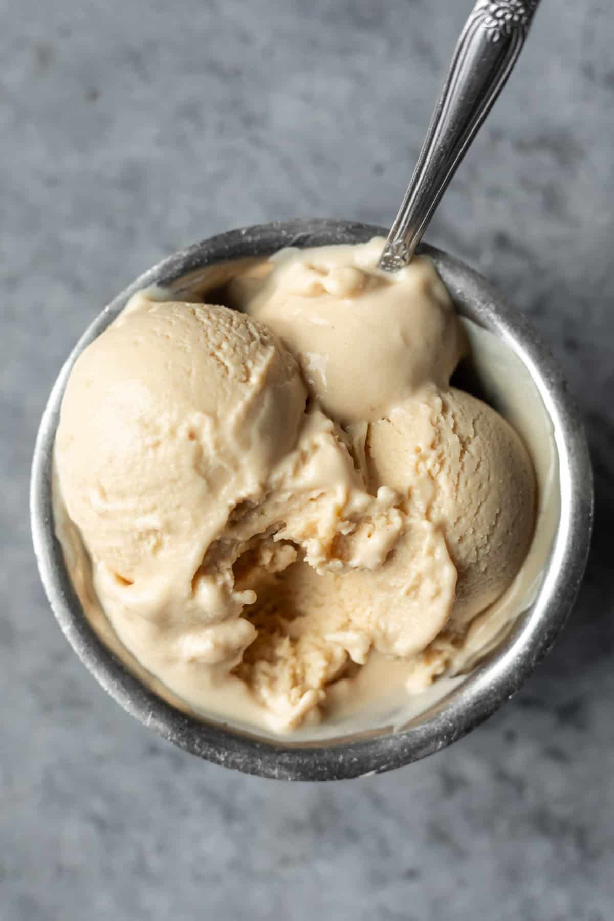 A scoop of vanilla ice cream in a metal cup with a metal spoon on a gray surface.