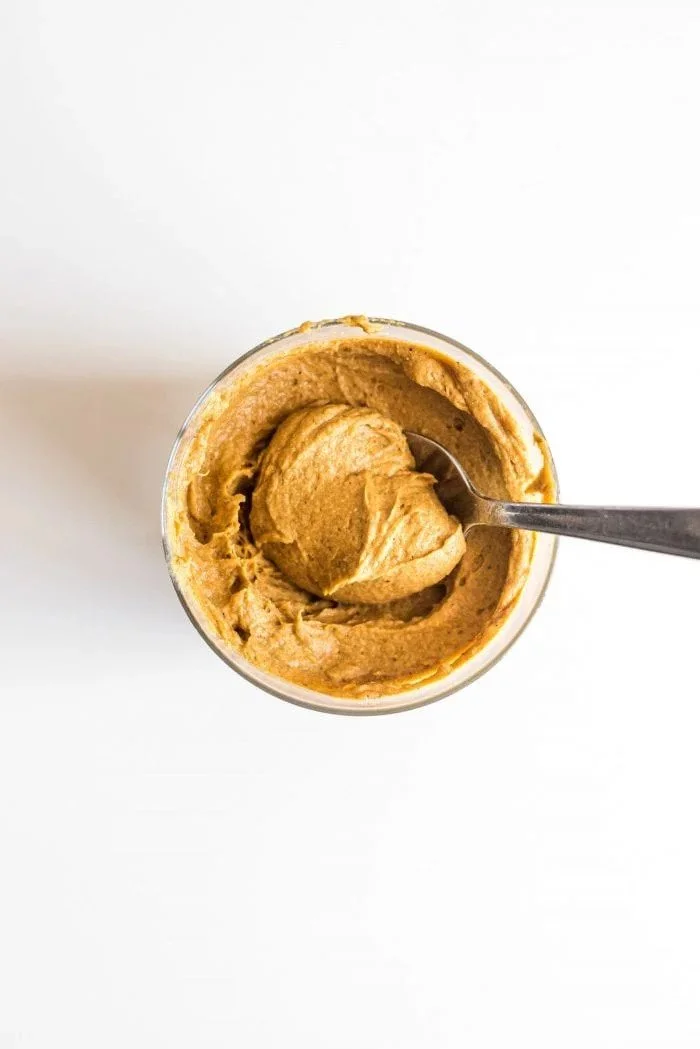 A glass jar filled with smooth peanut butter, with a metal spoon partially immersed in it, on a white background.