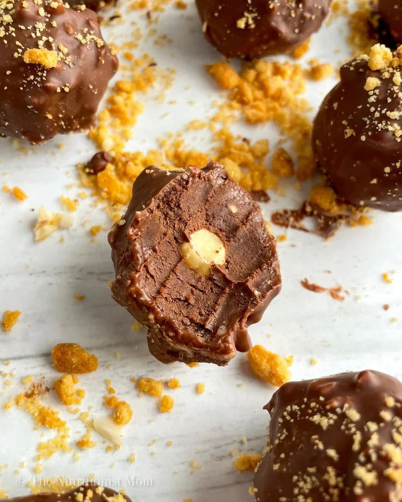 Close-up of chocolate truffles with a creamy filling and nut inside, sprinkled with cookie crumbs on a white surface.