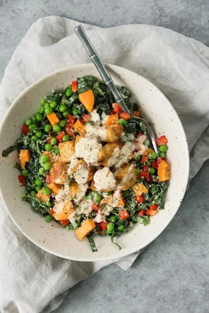 Bowl of cooked mixed vegetables including peas, diced carrots, red bell peppers, and greens, topped with tofu served with a fork.