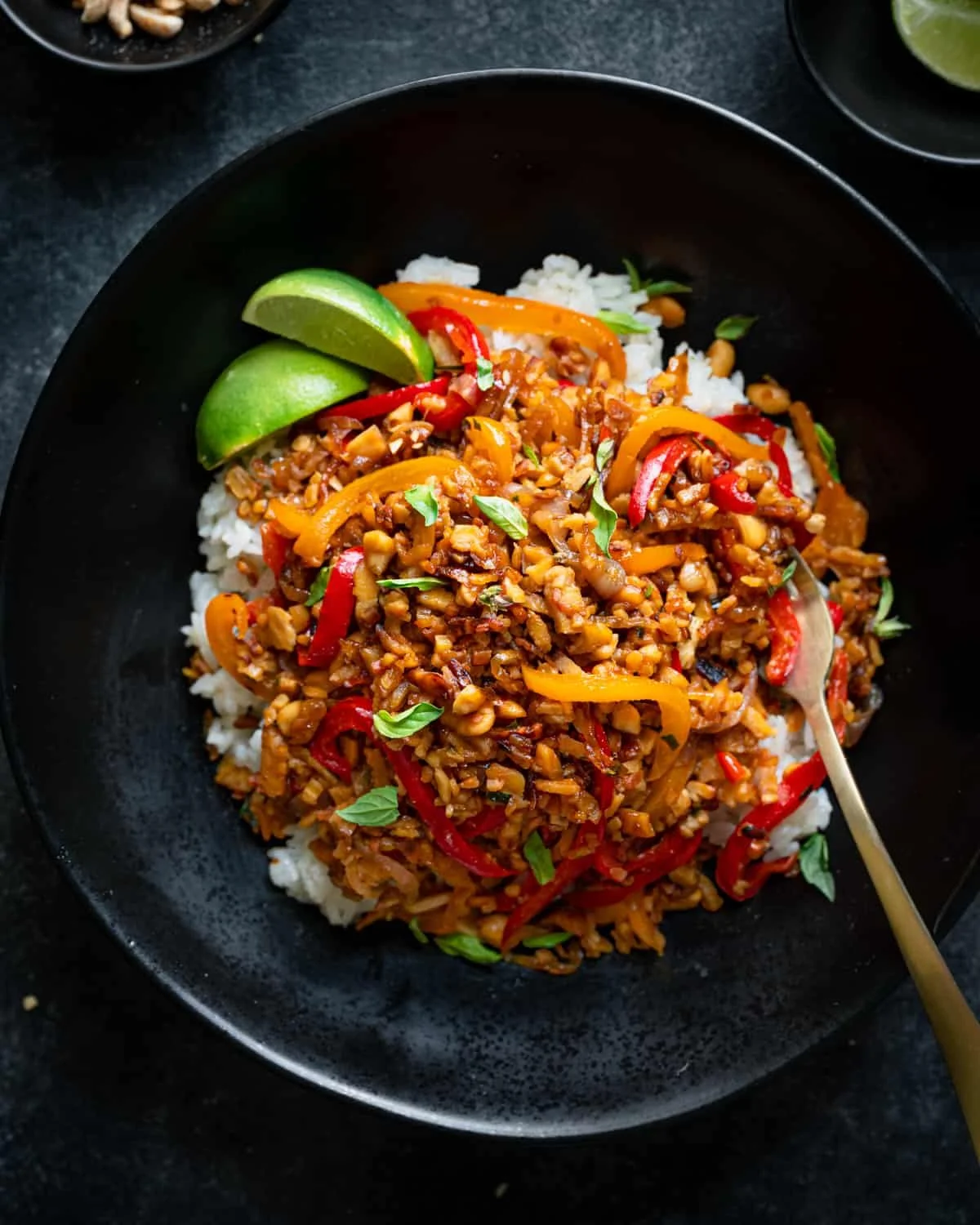 A black bowl filled with stir-fried vegan vegetables, red and yellow bell peppers, served over white rice, garnished with lime wedges and fresh basil leaves.
