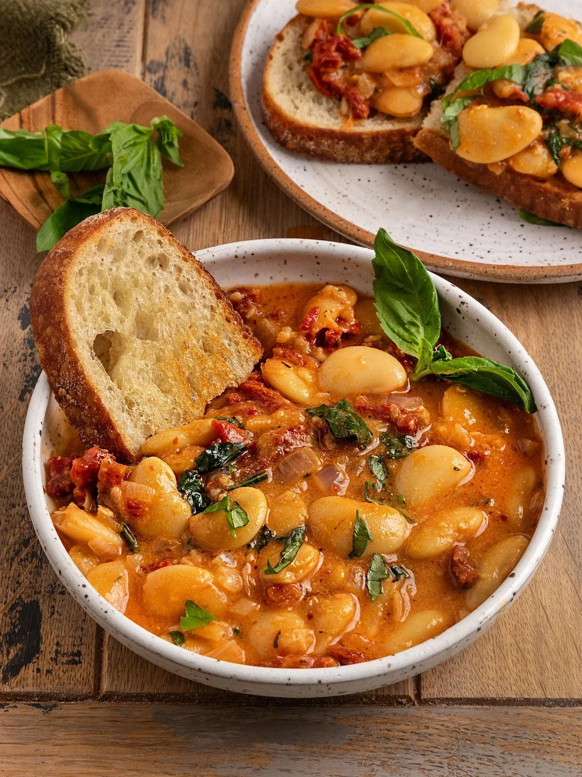 A bowl of white bean soup garnished with basil and served with a slice of toasted bread. In the background, two slices of bread topped with beans, herbs, and tomatoes on a plate.