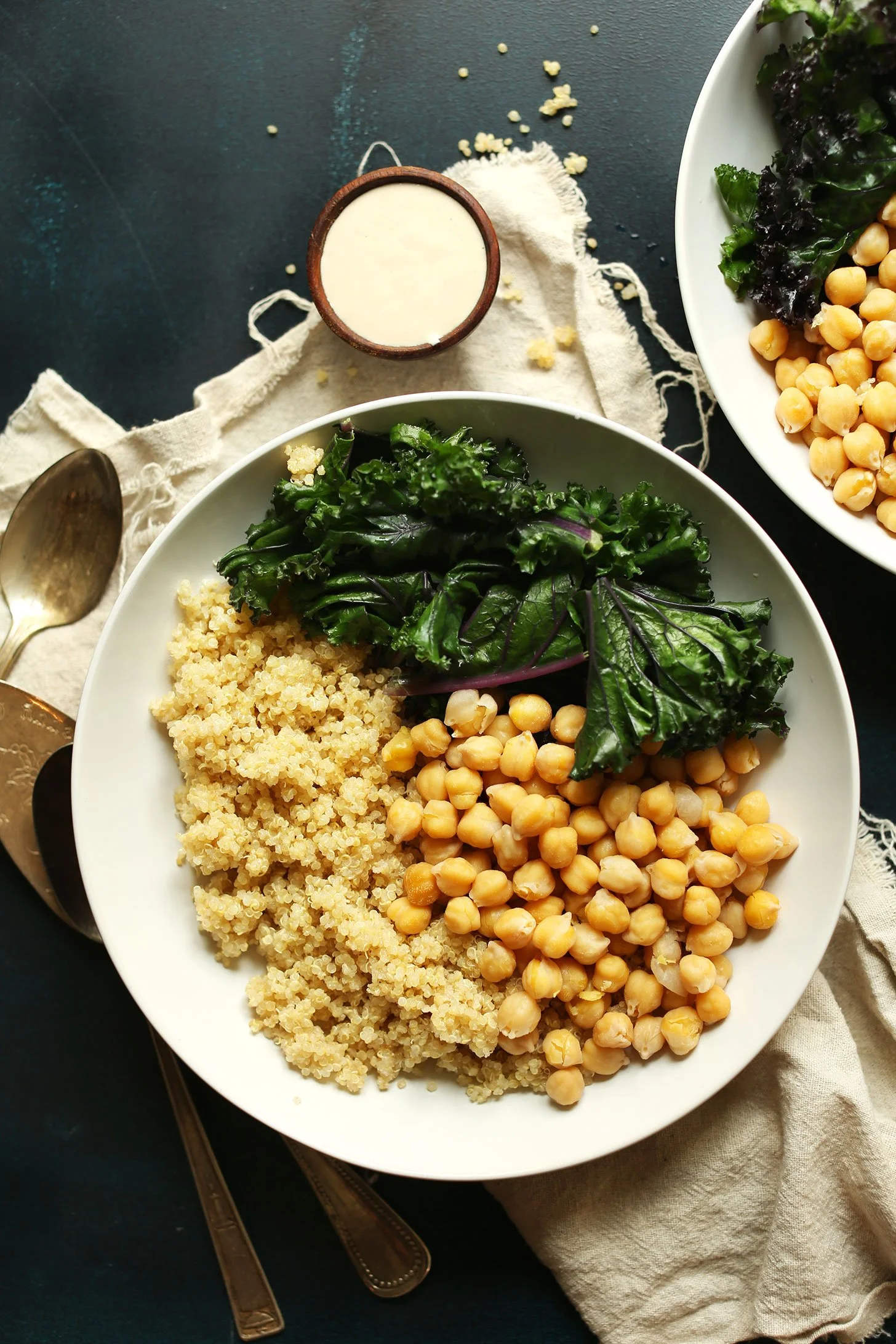 A white plate with a grain salad, chickpeas, and kale, accompanied by a small bowl of dressing or sauce and a second plate with more chickpeas and kale, set on a dark surface with a beige cloth, gold utensils, and scattered quinoa.